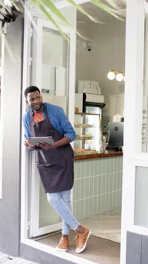 Vertical video: Standing at cafe entrance, male barista holding tablet and smiling confidently