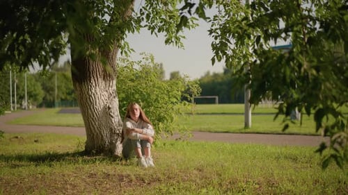 Thoughtful Woman Sitting Under Tree with Sunlit Face and Urban Stadium Background