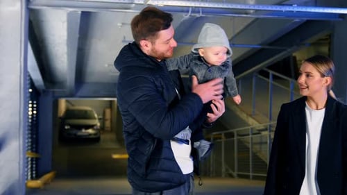 Happy family of three are in the car park. Man holding a baby kisses his son and waves him in hands.