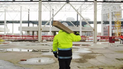 Rear View of Man Worker Walking Outdoors on Construction Site, Carrying Beam