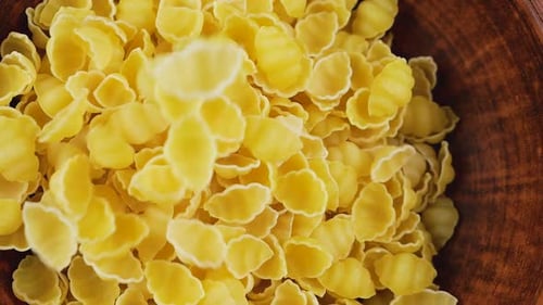 Shell-shaped pasta is poured into a brown ceramic bowl. Top view. Slow motion.