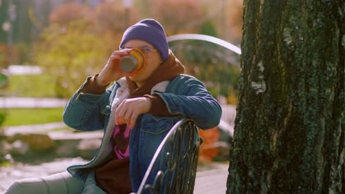 A Young Man with a Warm Hat is Drinking Coffee in the Park A Man in a Blue Denim Jacket and Glasses