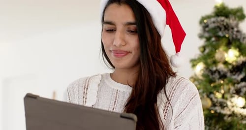 Mujer india con sombrero de Papá Noel usando una tableta cerca del árbol de Navidad, sonriendo calurosamente