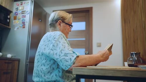Elderly Woman Reading Small Card at Kitchen Counter Magnets on Fridge Teapot on Table Quiet Domestic