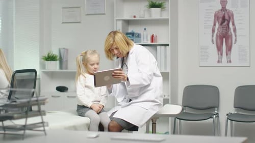 Female Doctor in Her Office Shows Tablet Computer to a Little Girl Patient.