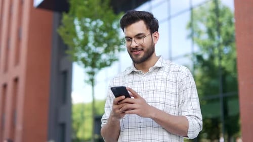 Handsome man is using a mobile phone while standing on the street near an modern building. Happy