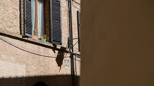 A sunlit street in Siena, Italy, with a window and shutters casting shadows on the brick wall
