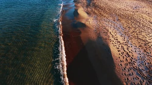 Soothing waves splash to the sandy shore. Beach covered with footsteps is lit by the setting sun.
