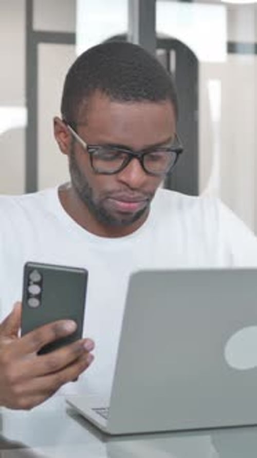 Young Man Using Phone and Laptop at Desk