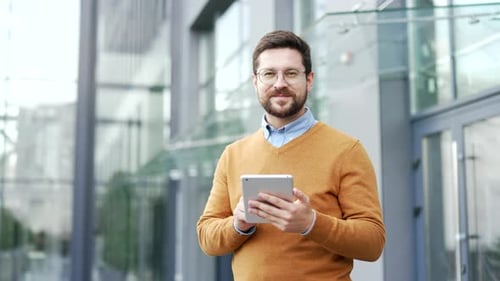Portrait of smiling businessman holding digital tablet standing on street near office building.