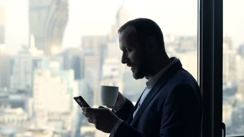 Businessman Drinking Beverage and Texting on Smartphone by Window in Office Adult