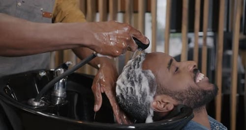 Smiling Male Client Getting Hair Wash with Water and Shampoo in Modern Barbershop
