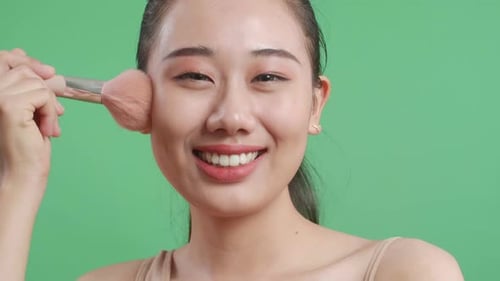 Close Up Of Beautiful Asian Woman Rouging Her Cheeks While Smiling To Camera On Green Screen
