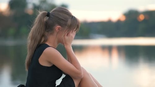 Upset Woman Sitting by Reflective Water at Dusk