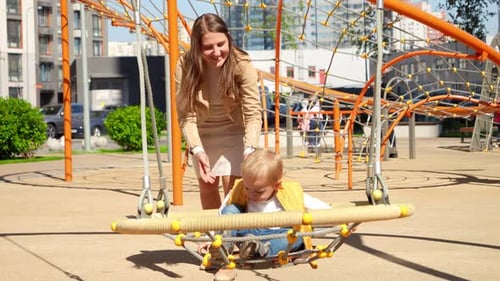 Happy smiling mother rocking her baby son in swing on the playground