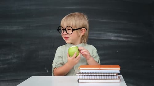 Back to School Happy Smiling Pupil in Round Glasses Sitting at the Desk Child Eating Green Apple in