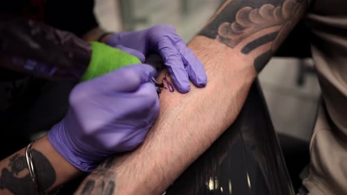 Un tatoueur professionnel portant des gants violets en nitrile inscrit méticuleusement le nom Belen sur un