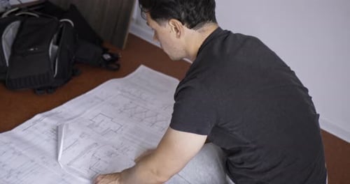 Work at Home on Weekends A Young Adult Man in Home Clothes Works with Drawings Laid Out on the Floor