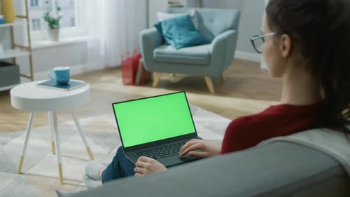 Young Woman at Home Works on a Laptop Computer with Green Mock-up Screen. She's Sitting On a Couch