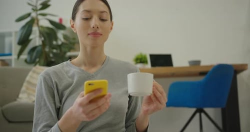 Smiling Woman Uses Phone While Holding Mug