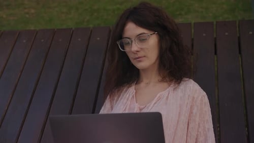 Handheld Close Up of a Curly Caucasian Woman Working on Her Laptop Computer on a Park Bench