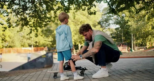 Dad Helps His Little Son Put on Protective Clothing Before Riding a Skateboard at the Skatepark in