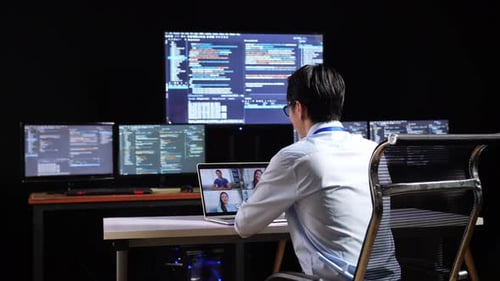 Asian Male Programmer Having A Meeting By A Laptop While Writing Code On Multiple Monitors