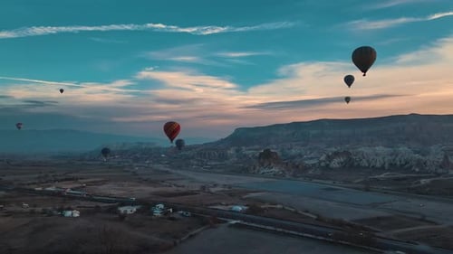 Drone view of numerous hot air balloons flying low over the ground at sunrise in Cappadocia, with mo