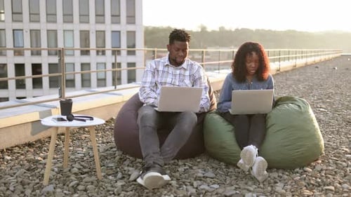 African Couple Using Laptops While Sitting in Beanbags at Rooftop of Office