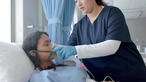 Diverse female doctor putting oxygen mask on female patient in hospital bed, slow motion