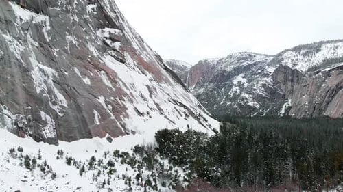 Aerial drone footage slowly flying past snowy granite cliffs towering over Yosemite valley in winter