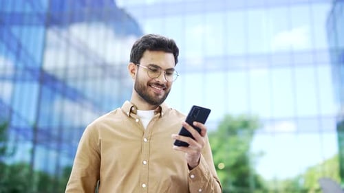 Happy businessman standing near modern glass business office building holding smartphone in hands.