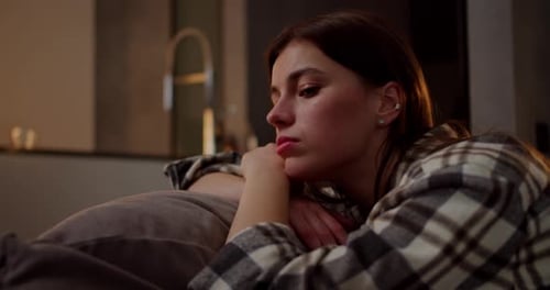 A Calm and Bored Brunette Girl in a Checkered Shirt Leans on a Pillow on a Gray Sofa She is Bored