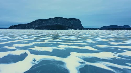 Aerial footage flying low over a frozen lake with snowy patterns towards an isolated mountain with a