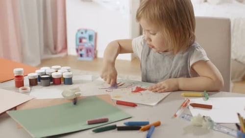 Симпатичная маленькая девочка сидит за своим столом и рисует карандашами. Ее комната розовая и уютная.