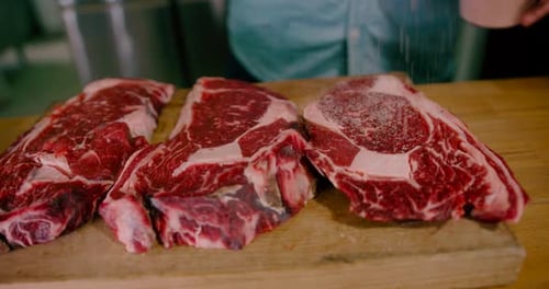 Close up view of raw beef steaks on the table while adding salt with pepper and spices at fresh meat