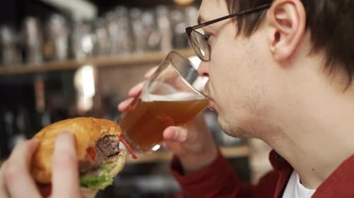 Young Man Leisurely Eating Burger Drinking Beer at Casual Bistro Enjoying Laidback Meal with Hearty