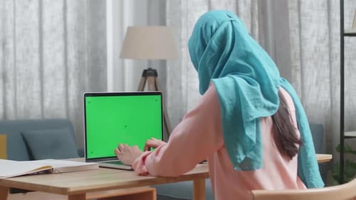 Back View Of A Muslim Woman With A Book On The Table Typing On Mock Up Green Screen Laptop