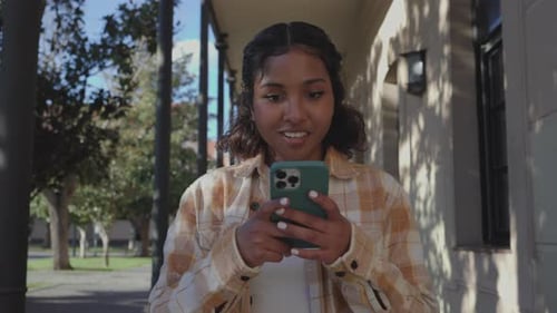 Student walking on university campus, smiling while messaging on smartphone, enjoying digital commun