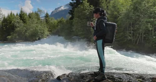 Female Traveler with a Backpack Drinking Water in Nature in the Forest Near a Mountain River
