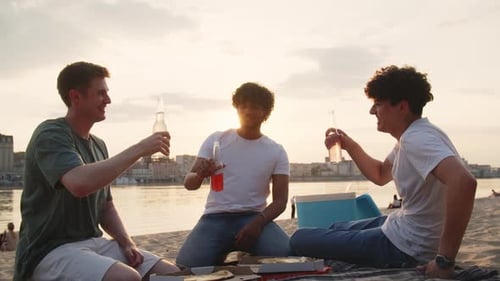 Three Young Men Friends on a Summer Evening Sit on the Beach at Sunset Joyfully Celebrate the