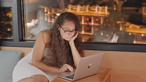 Relaxed Woman with Glasses and City View Casual Woman in Hotel Room Browsing Computer While City