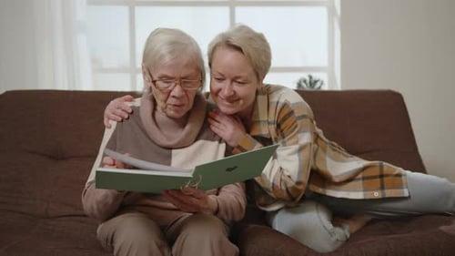Hija amorosa con madre anciana mira el álbum de fotos en un abrazo, disfrutando de los recuerdos