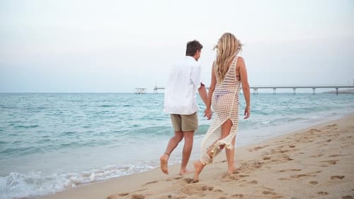 Young Couple Joyfully Walking Hand in Hand Along the Sandy Beach at Sunset Enjoying a Romantic