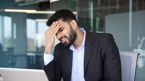 Unhealthy businessman suffering from headache sitting at workplace in office. Unwell worker holding