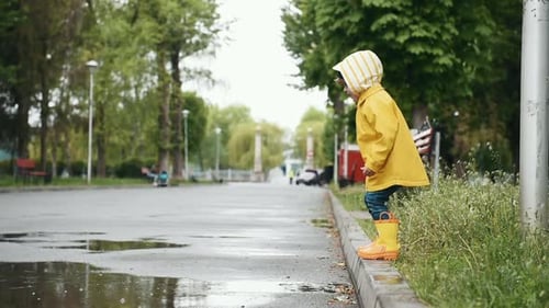 Kid in yellow waterproof cloak and boots jumping into puddle outdoors after the rain