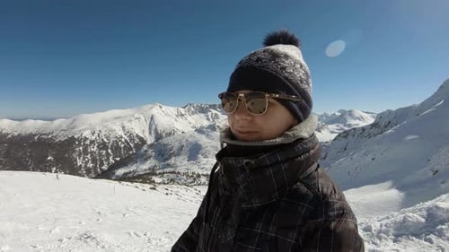 Girl with winter hat and sunglasses, looking and smiling to the camera at a top of a mountain