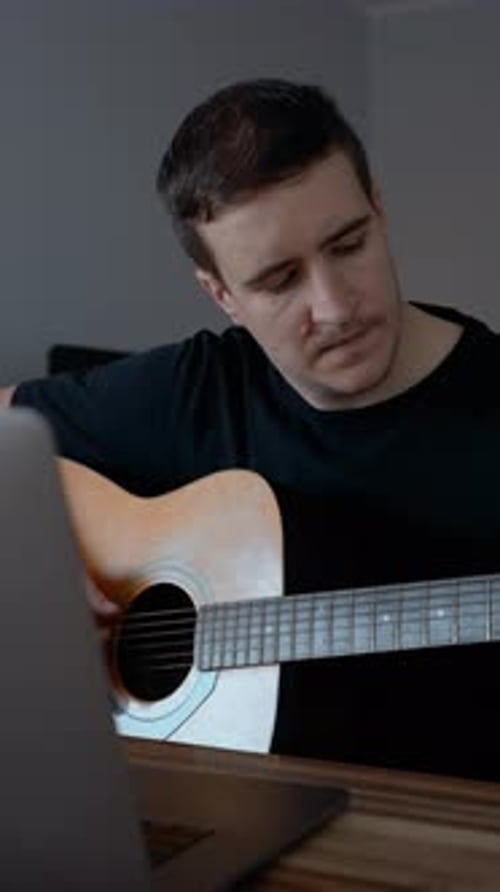 Man Plays Acoustic Guitar at Home with Laptop