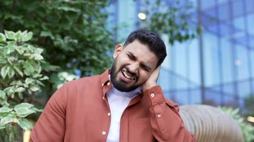 Sick businessman suffers from ear pain sitting on a bench on the street. Upset male touches his ear