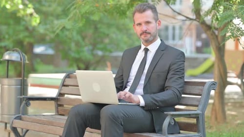 Thumbs Down by Middle Aged Businessman Using Laptop Outdoor on Bench